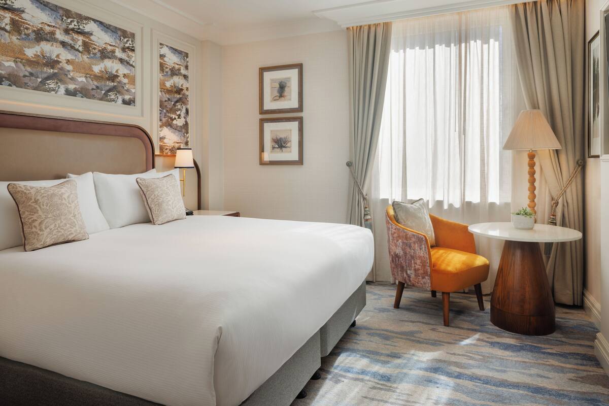 A hotel room with a large bed, a chair, a small table, and artwork on the walls.