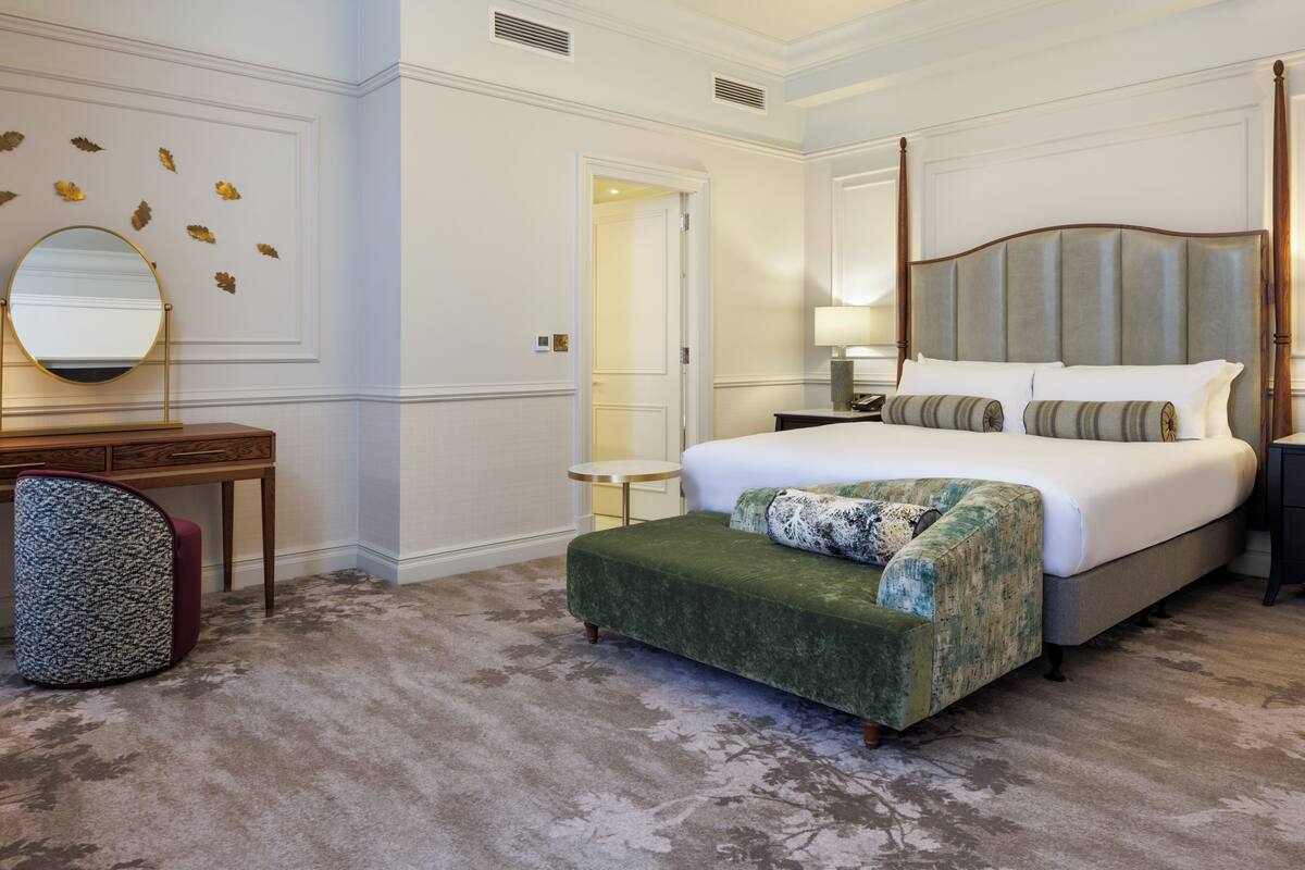 A hotel room with a large bed, a desk with a chair, a round mirror, and a small table.