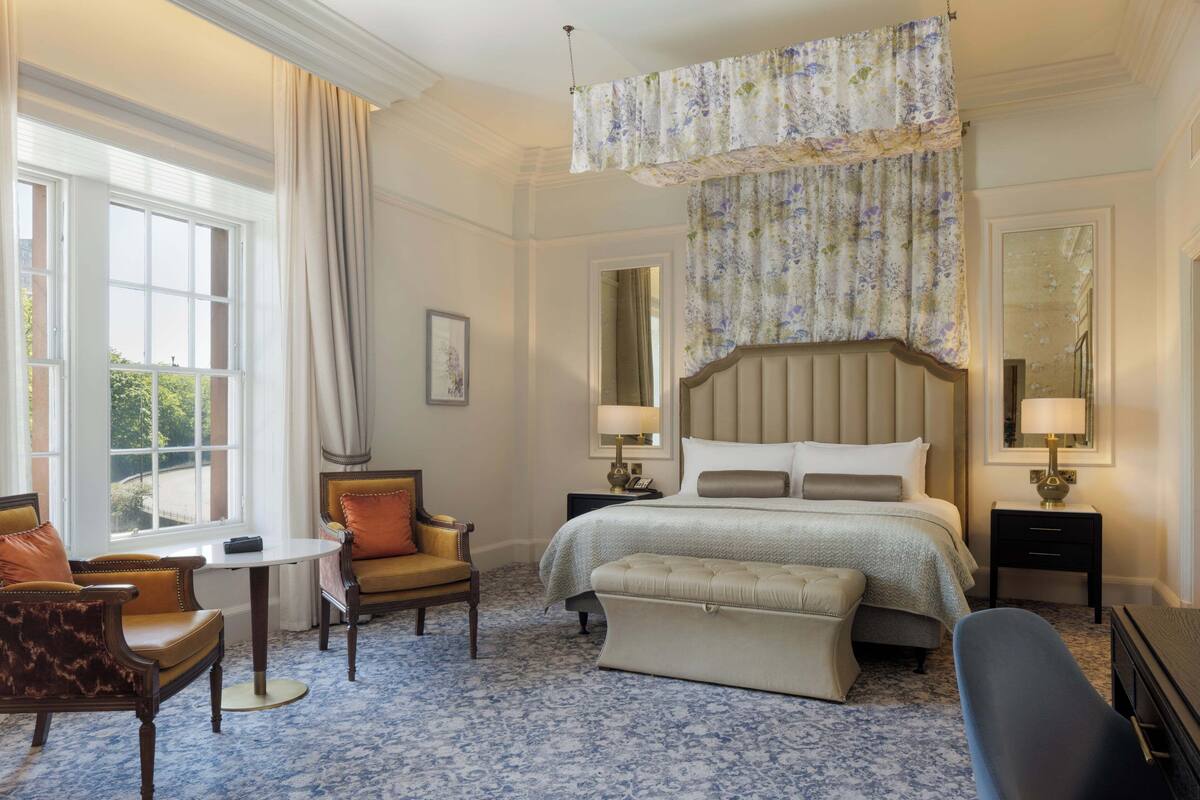 A hotel room with a large bed, a sitting area with a table and chairs, and a view of the outdoors through a window.