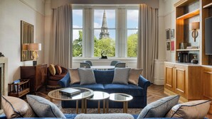 A living room with a blue sofa, two round coffee tables, and a view of a church steeple through large windows. - The Caledonian Edinburgh, Curio Collection by Hilton (Edinburgh)