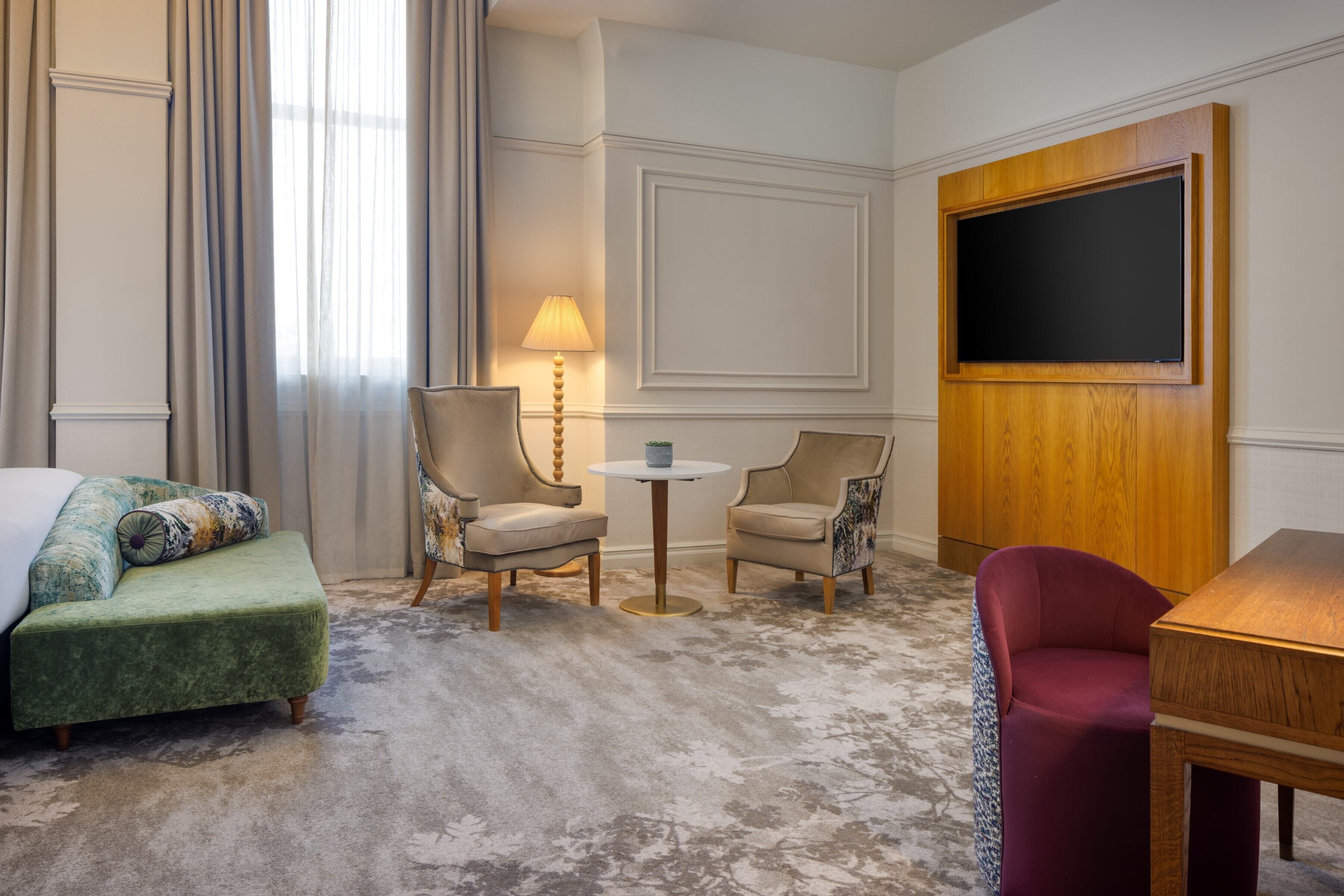 Een hotelkamer met een groene bank, twee fauteuils, een kleine ronde tafel, een rode stoel en een houten kast met een flatscreen televisie.
