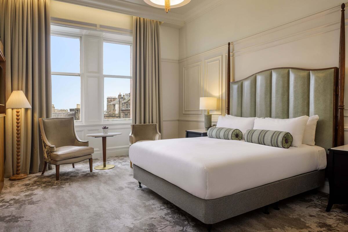 A large, neatly made bed with a headboard, two bedside tables, a chair, and a small round table.