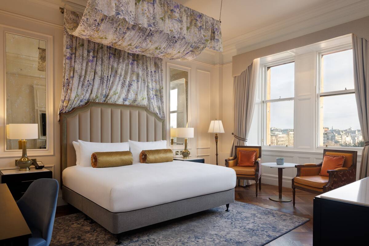 A hotel room with a large bed, two bedside tables with lamps, a small round table, two armchairs, and a view of the city through large windows.