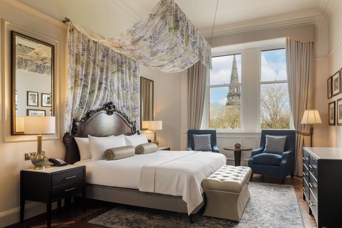 A spacious bedroom with a large bed, a sitting area with two armchairs, a small round table, and a view of a tower through the window.
