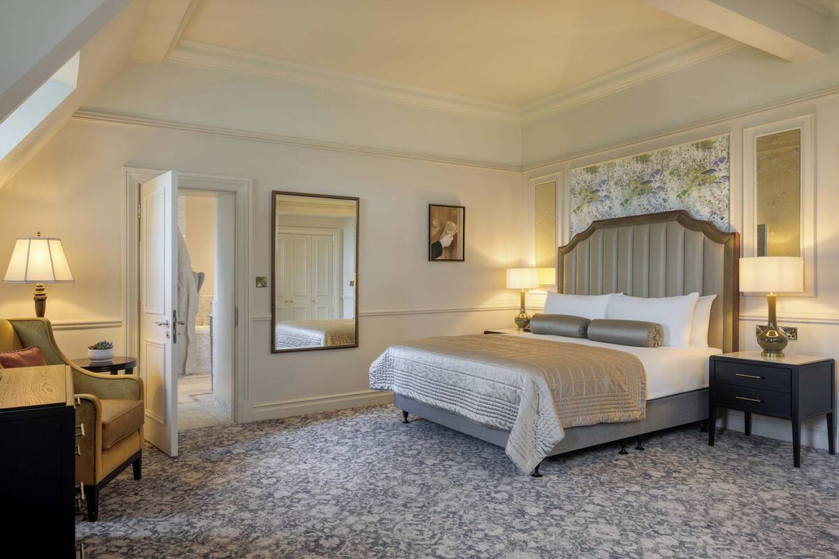 A bedroom with a large bed, two bedside tables, a chair, a mirror, and a door.