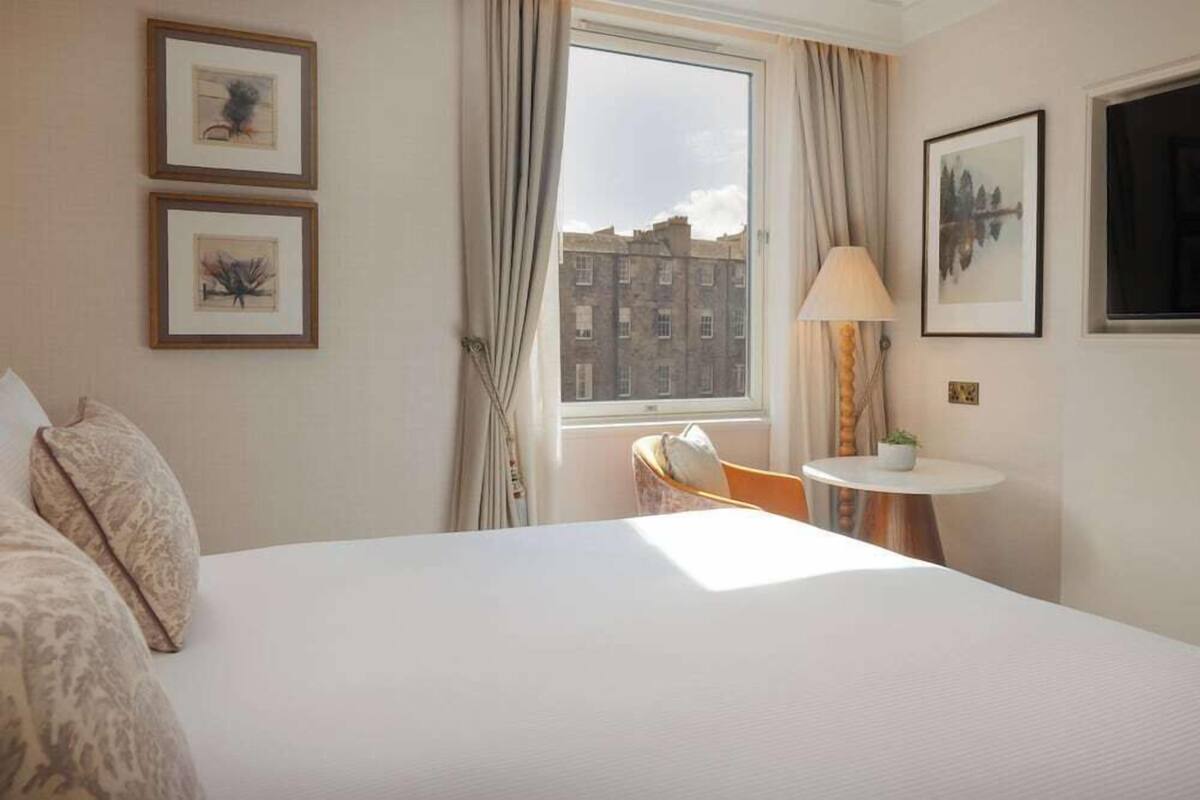 A hotel room with a bed, a chair, a small table with a lamp, and two framed pictures on the wall.