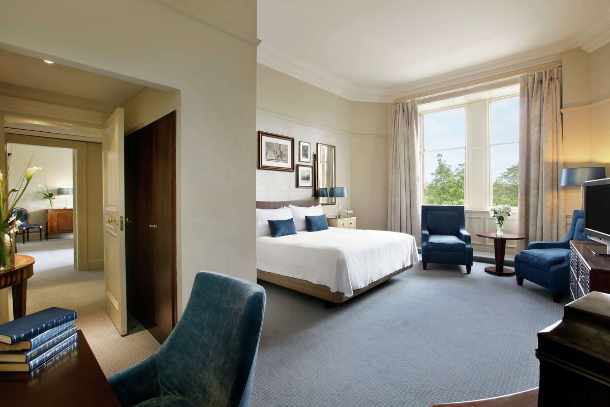 A hotel room with a bed, two armchairs, a small table, a TV, and a view of the outdoors.