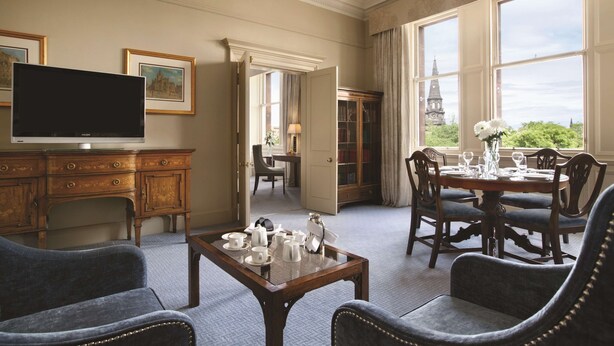 A spacious room with a large television, a dining area with a table set for four, and a view of a clock tower through the windows. - The Caledonian Edinburgh, Curio Collection by Hilton (Edinburgh)