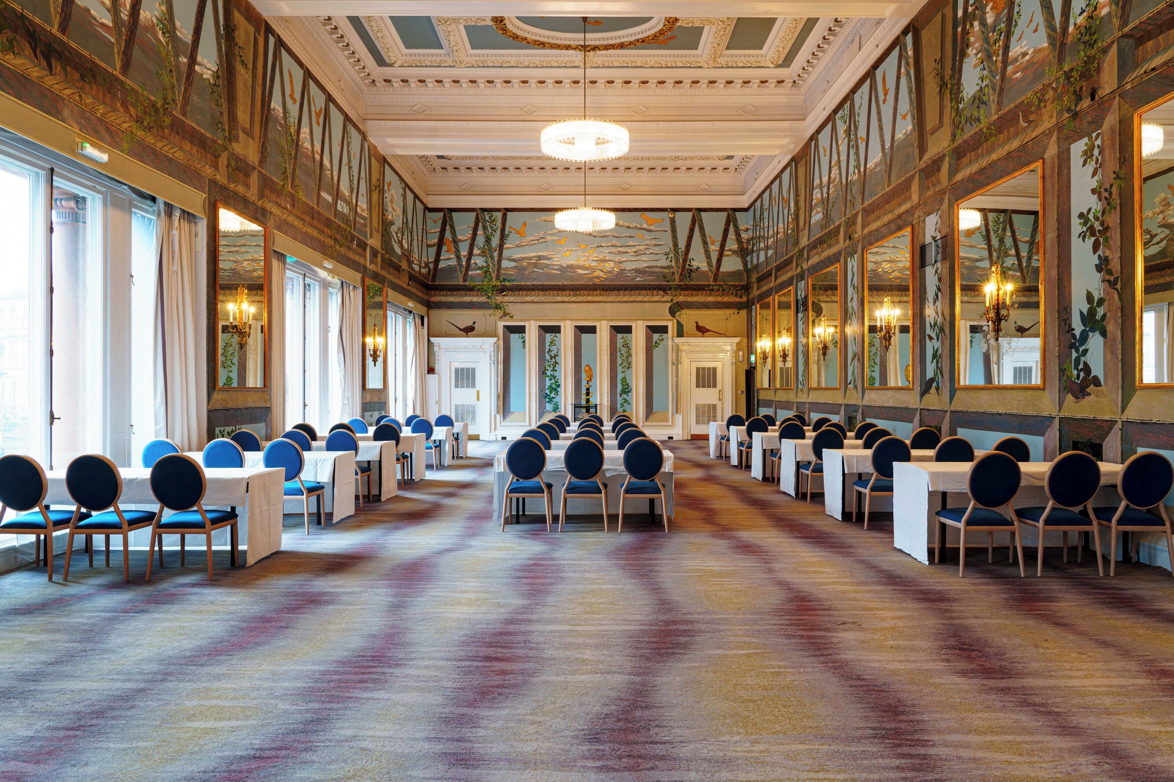 A large hall with rows of tables and chairs, a patterned carpet, and ornate ceiling decorations.