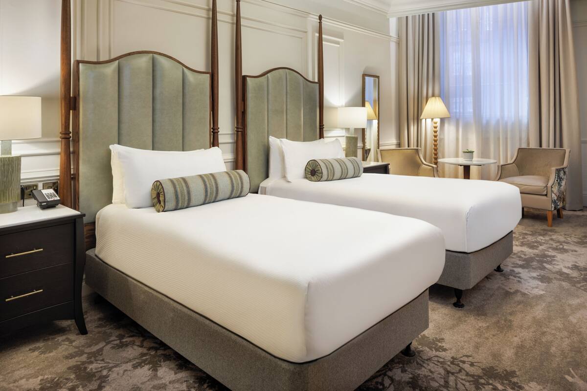 A hotel room with two beds, a nightstand, a chair, a small table, and a lamp.