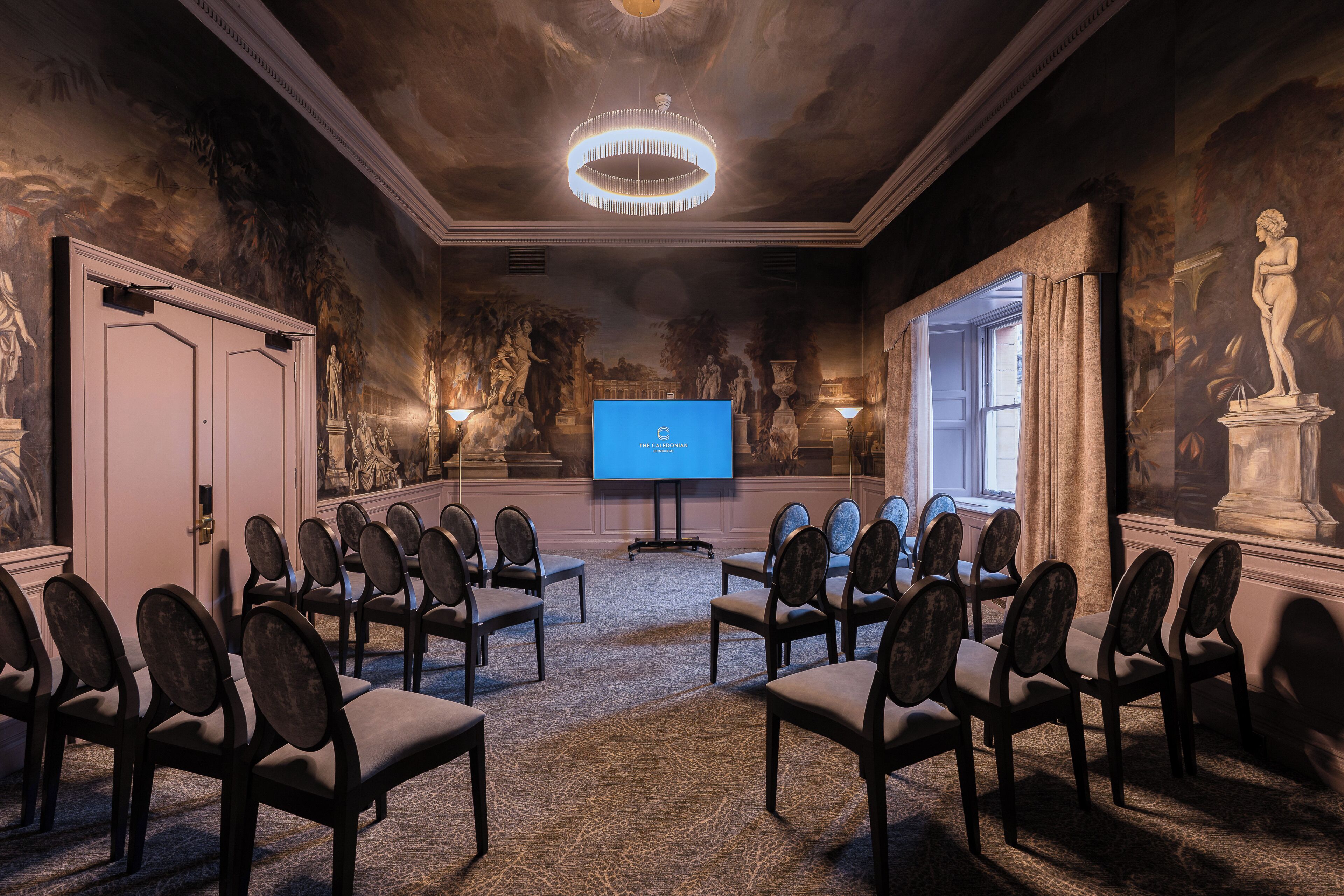 A conference room with a large screen, rows of chairs, and classical artwork on the walls.