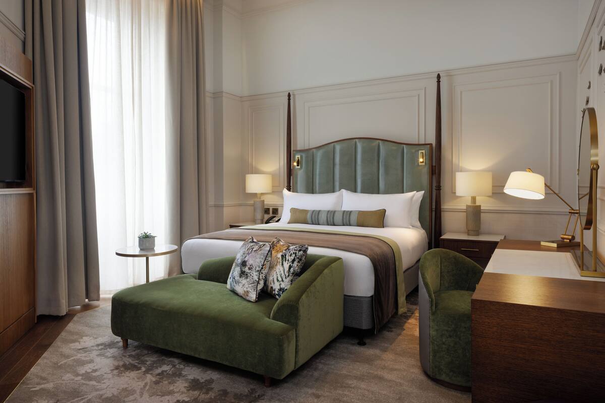 A hotel room with a large bed, a green upholstered sofa, a wooden desk, and a small side table with a lamp.