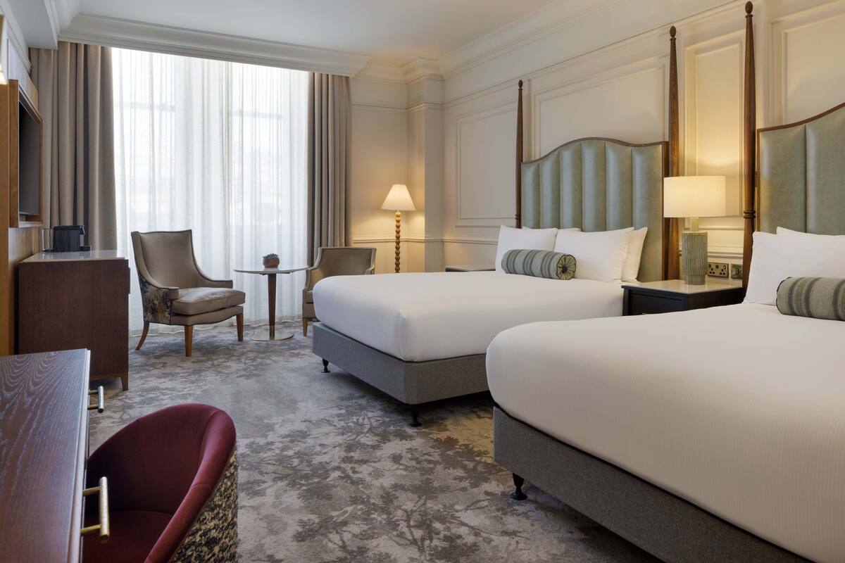 A hotel room with two beds, a desk, a chair, and a lamp.