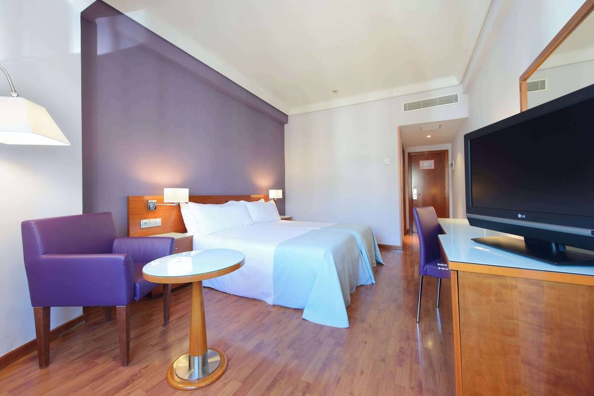 Ein Hotelzimmer mit einem Bett, einem Fernseher, einem violetten Sessel, einem kleinen Tisch und einem Holzbüro.
