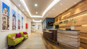 Un hall d'hôtel avec un comptoir d'accueil en bois, un téléviseur et un panneau indiquant « Enregistrement prioritaire ». On y trouve trois canapés verts avec des coussins roses, et un escalier en arrière-plan.