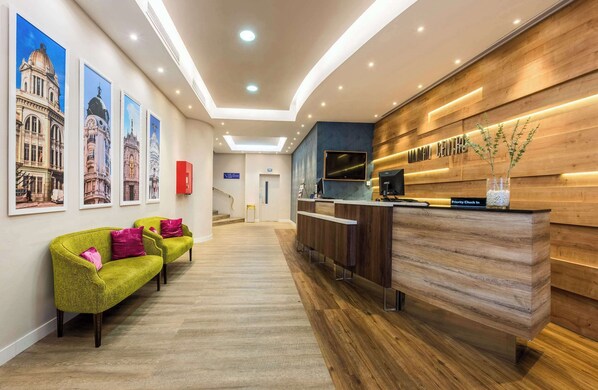 Zona de recepción de hotel con un mostrador de madera, un televisor y un letrero que dice 'Priority Check In'. Hay tres sofás verdes con cojines rosas y una escalera al fondo.
