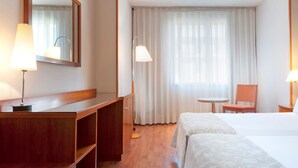 Une chambre d’hôtel avec un bureau en bois, deux tables de chevet, un lit recouvert d’une couvre-lit à motifs, une chaise, une petite table ronde et une grande fenêtre avec des rideaux légers.