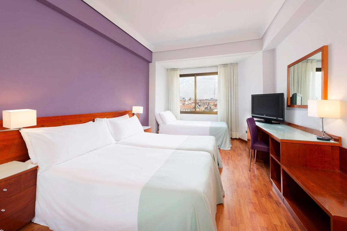 Ein Hotelzimmer mit zwei Betten, einem Schreibtisch, einem Fernseher und einem Fenster mit Blick auf die Stadt.