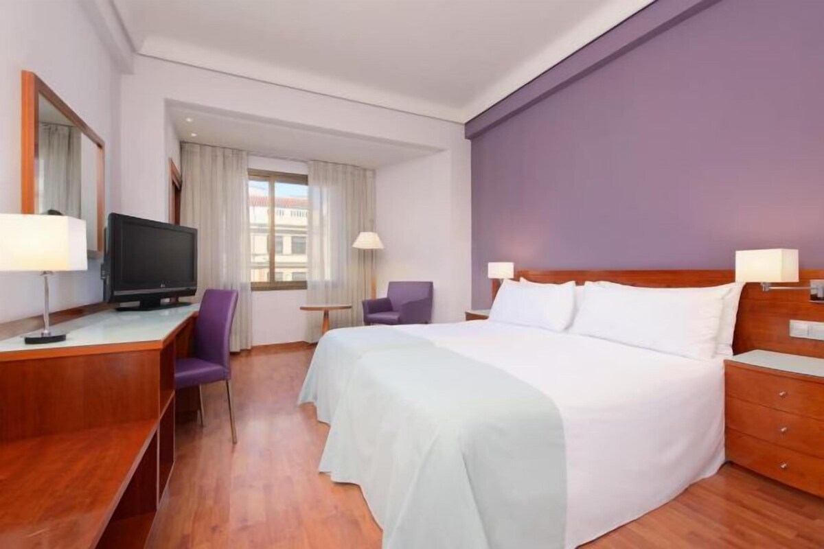 Een hotelkamer met een bed, een bureau met een televisie, een stoel en een raam met gordijnen.