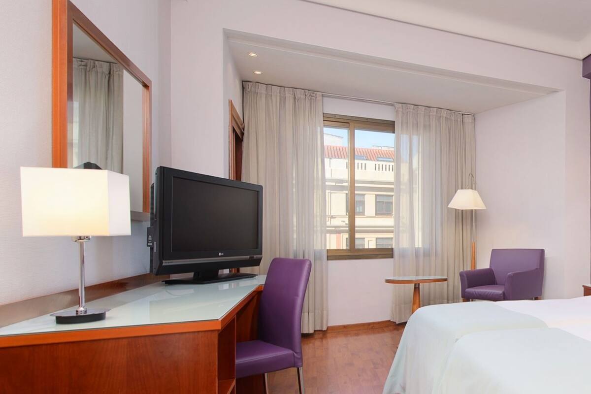 Ein Hotelzimmer mit einem Holztisch, einem Fernseher, einem violetten Sessel, einem Bett, einem kleinen Tisch und einem Fenster mit Vorhängen.