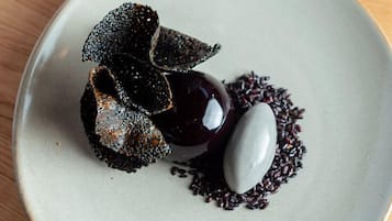 Un plato con un postre de color oscuro, posiblemente un mousse de chocolate, acompañado por un objeto esférico blanco y una guarnición oscura y texturizada.