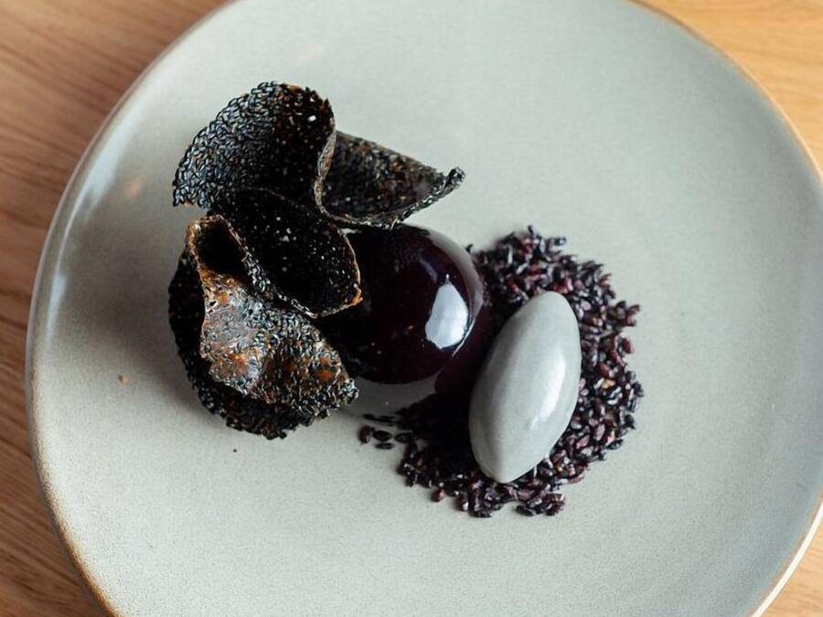 Un plato con un postre de color oscuro, posiblemente un mousse de chocolate, acompañado por un objeto esférico blanco y una guarnición oscura y texturizada.