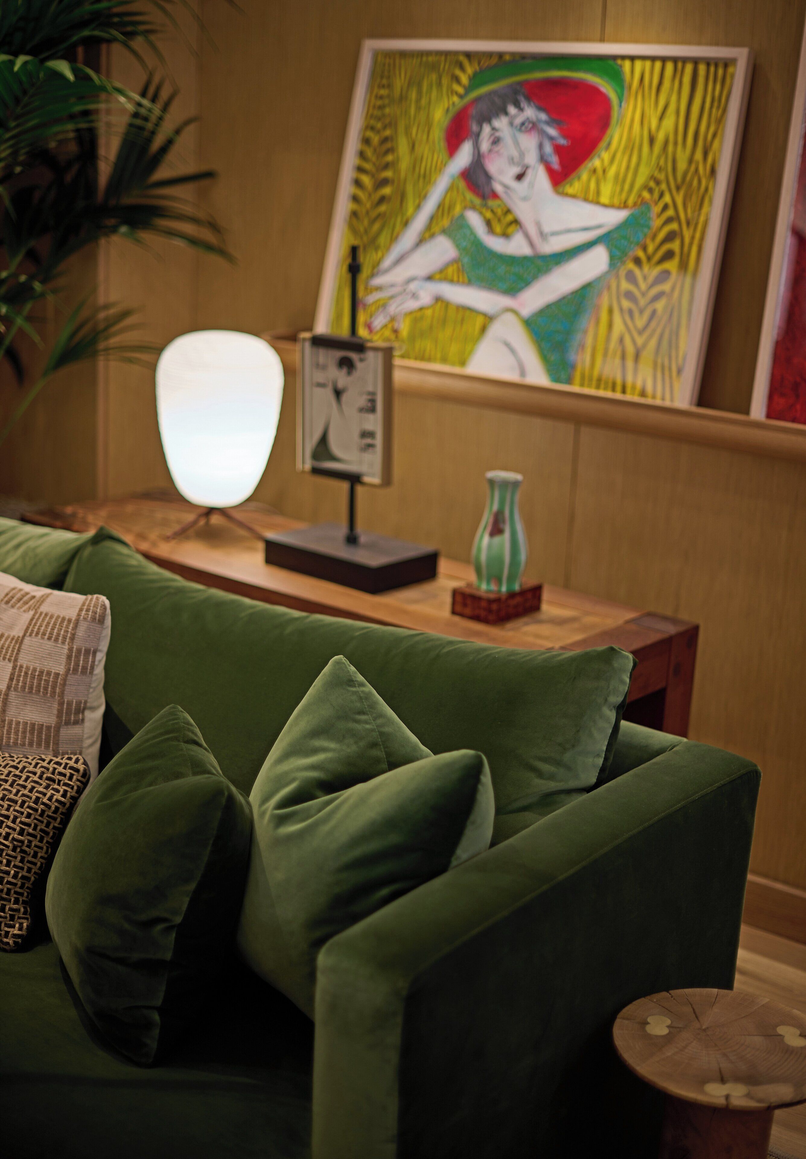 A green sofa with multiple cushions, a wooden side table with a vase, and a framed painting on the wall.