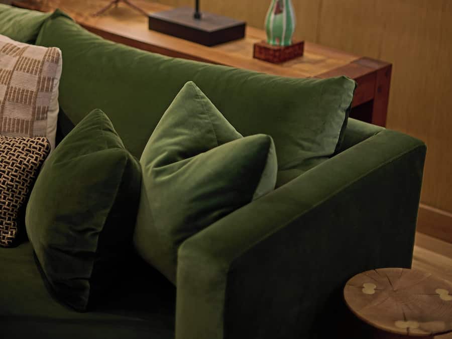 Un sofá verde con varios cojines, una mesita de madera con un florero y un cuadro en la pared.
