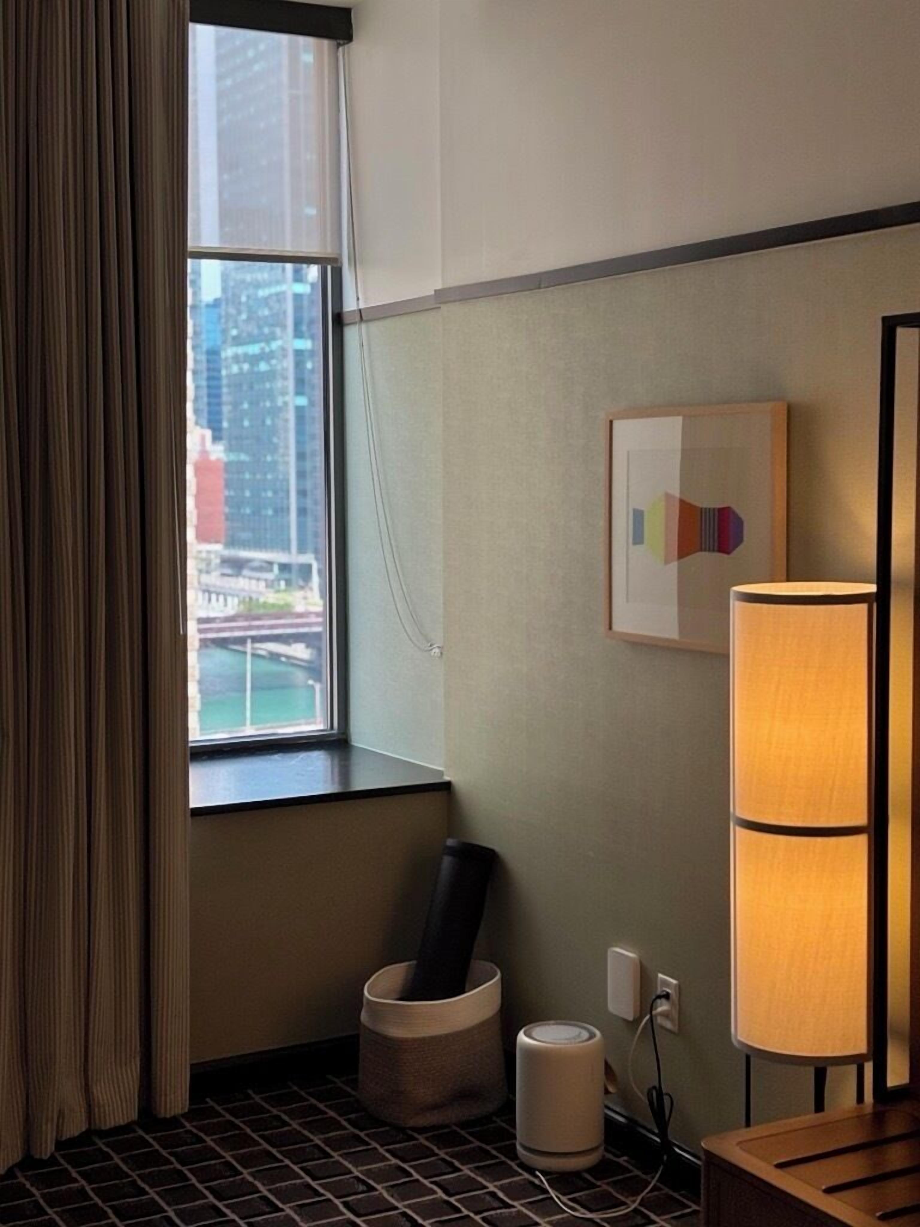A hotel room with a large window, a city view, a framed artwork, a lamp, and a trash bin.
