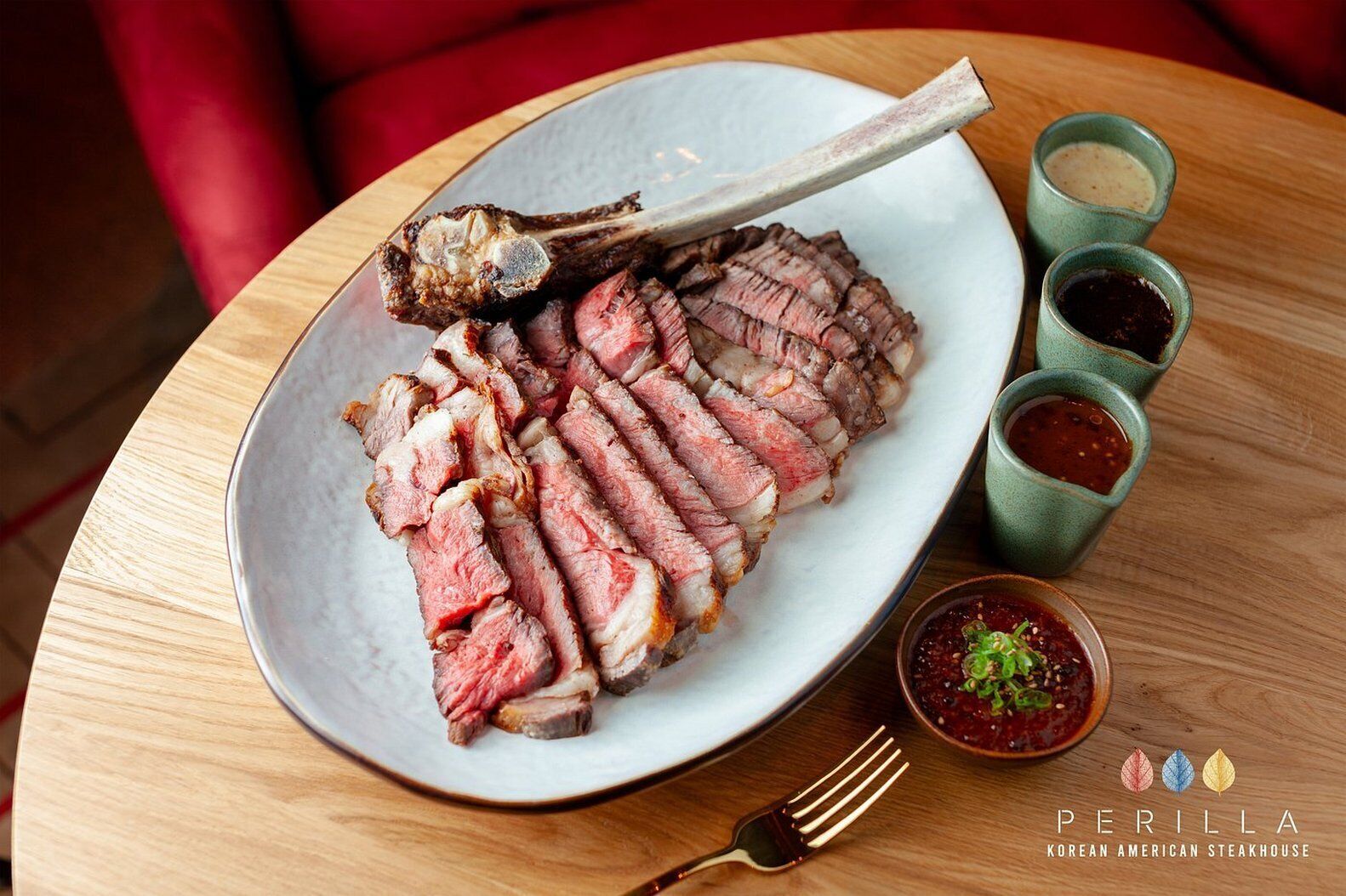 A plate of sliced steak with bone, accompanied by three small bowls of sauces, on a wooden table.