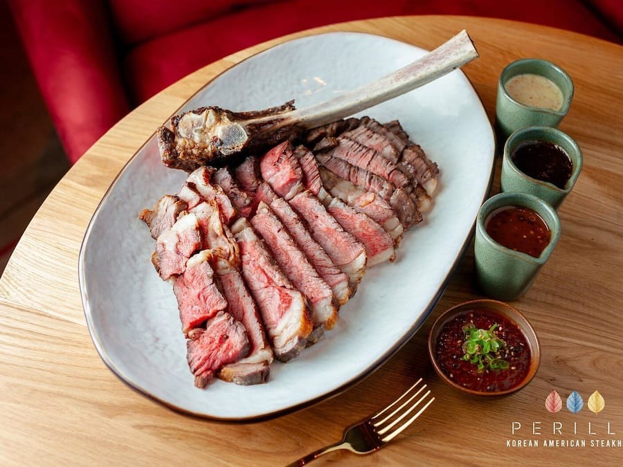 Un plato de carne en bistec en rodajas, con hueso, acompañado de tres recipientes pequeños con salsas, sobre una mesa de madera.