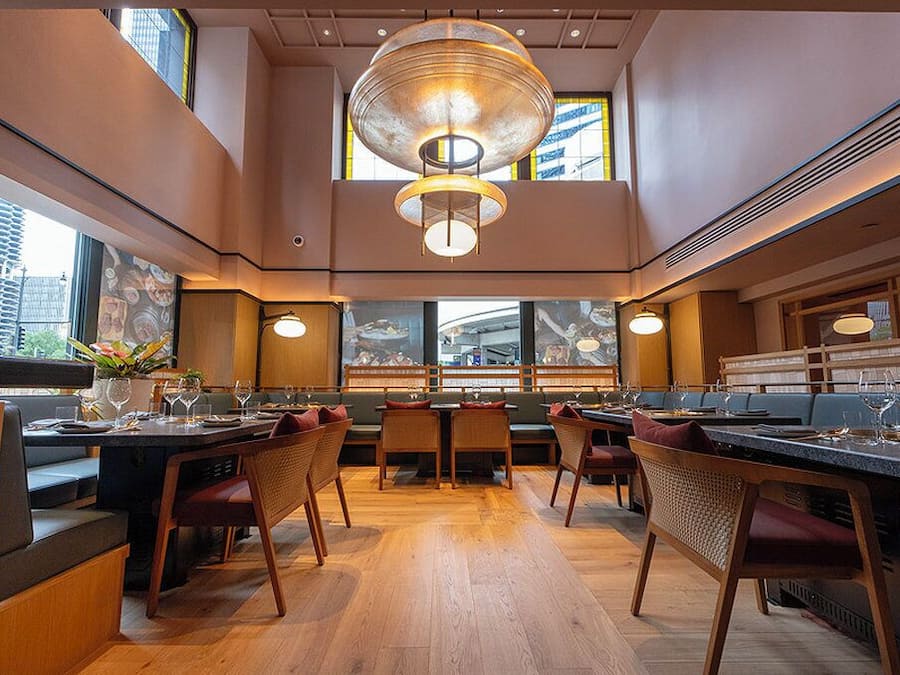 A modern restaurant with high ceilings, wooden flooring, and a central chandelier. Tables are set with wine glasses and red cushions.