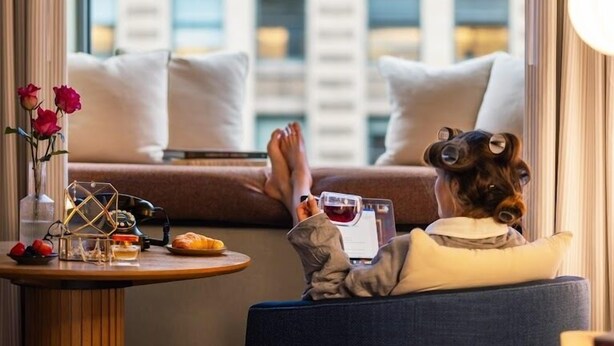Une personne se détend sur un canapé avec un ordinateur portable, un verre de vin rouge et une petite table sur laquelle se trouvent un vase de fleurs et un téléphone.