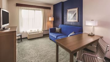 A hotel room with a blue sofa, a wooden table, a TV, a lamp, and a framed picture on the wall.