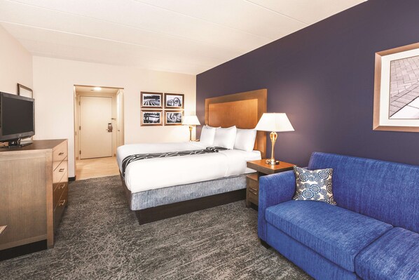 Habitación de hotel con una cama, un sofá azul, un escritorio con televisor y obras de arte enmarcadas en las paredes.