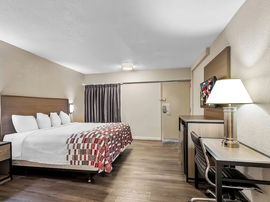 A hotel room with a bed, bedside table, lamp, desk, chair, and a door with a curtain.