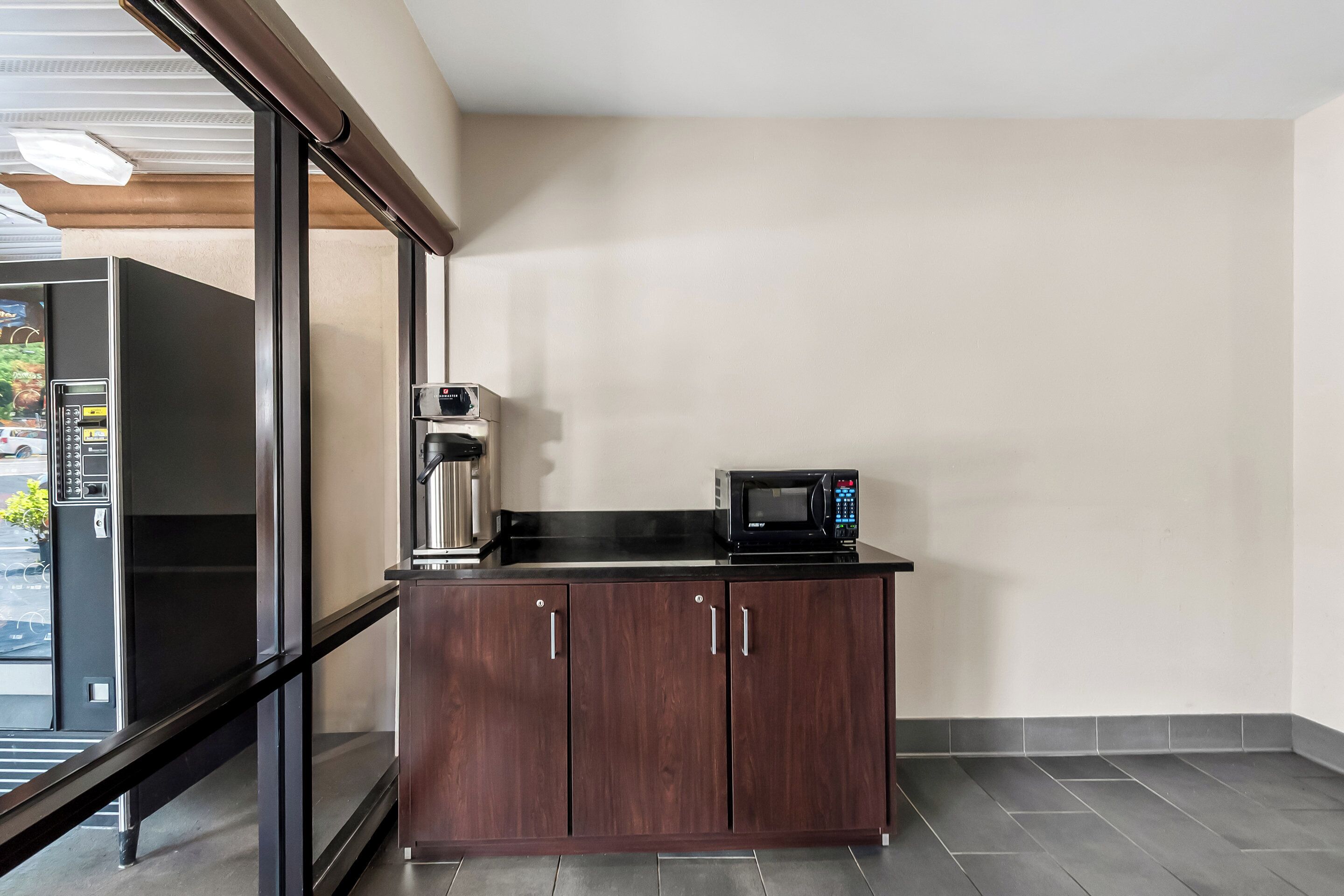 Una kitchenette con microondas y cafetera sobre una encimera oscura, un refrigerador y un vidrio corredizo que da a un espacio exterior.
