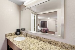 A bathroom with a granite countertop, a white sink, a coffee maker, and a mirror reflecting a room with a bed and curtains. - Red Roof Inn Washington, DC (Washington)