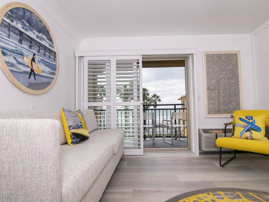 A living room with a sofa, a yellow armchair, a painting of a surfer, and a view through a glass door.