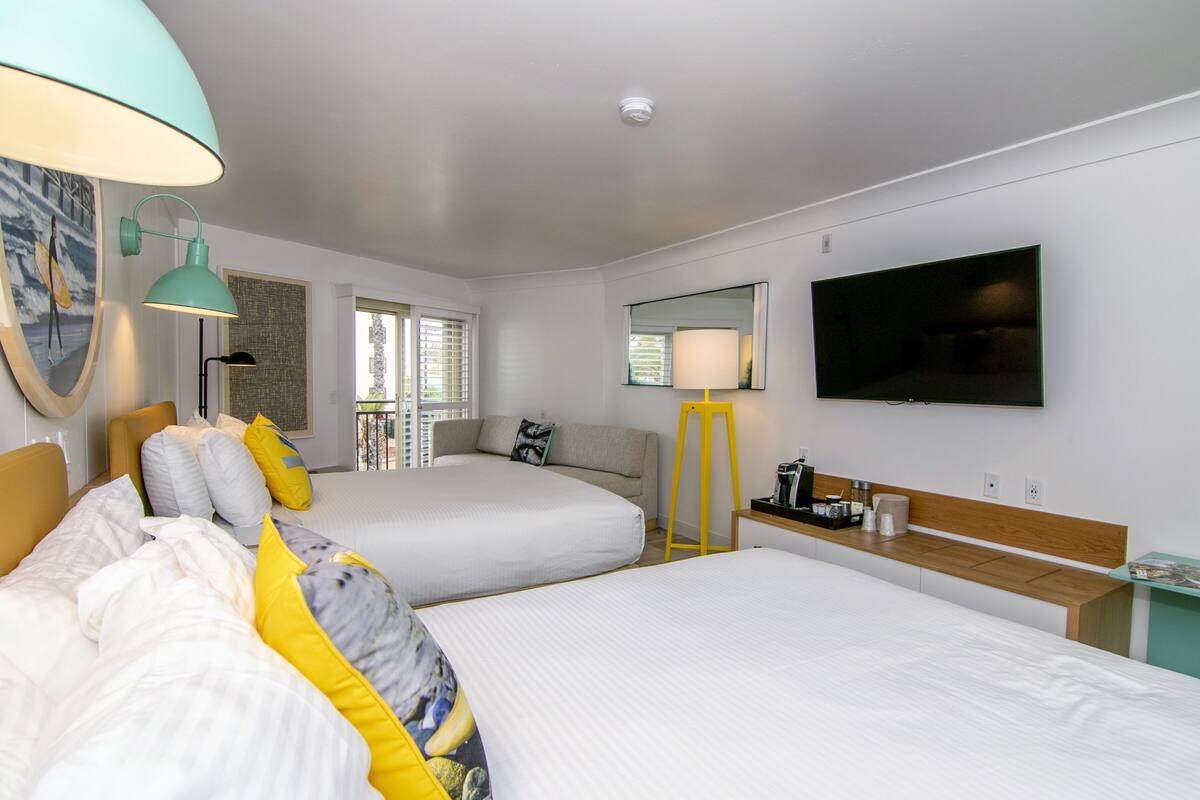 A hotel room with two beds, a TV, a desk with a lamp, and a window with blinds.