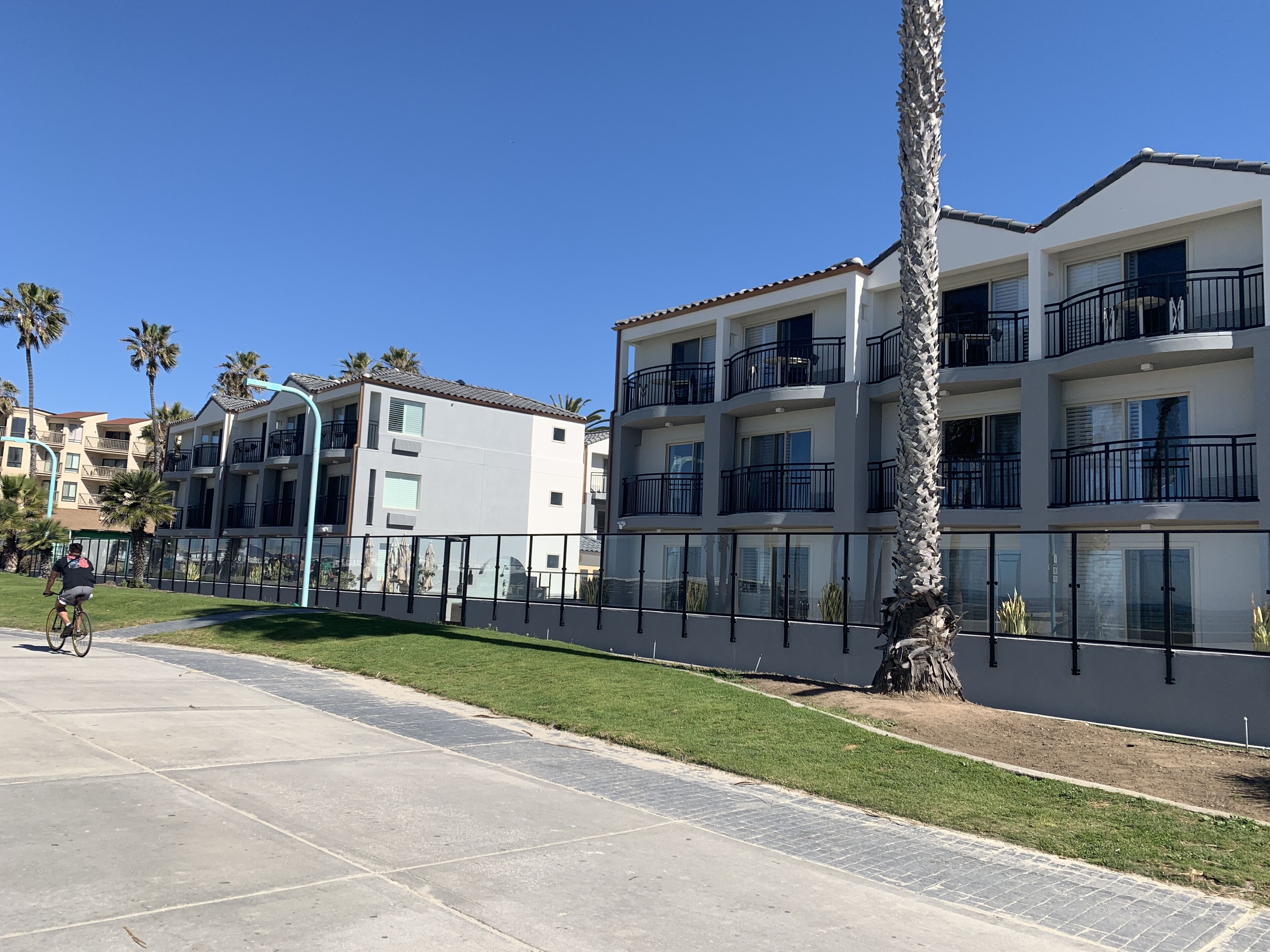 A multi-story residential building with balconies, a paved walkway, and palm trees.