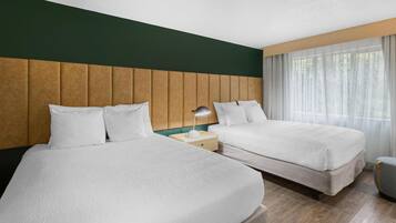 A hotel room with two beds, a wooden headboard, a bedside table with a lamp, and a window with sheer curtains.