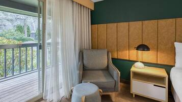 Camera d'albergo con vista sul verde circostante, una poltrona grigia, un comodino con una lampada e un piccolo poggiapiedi.