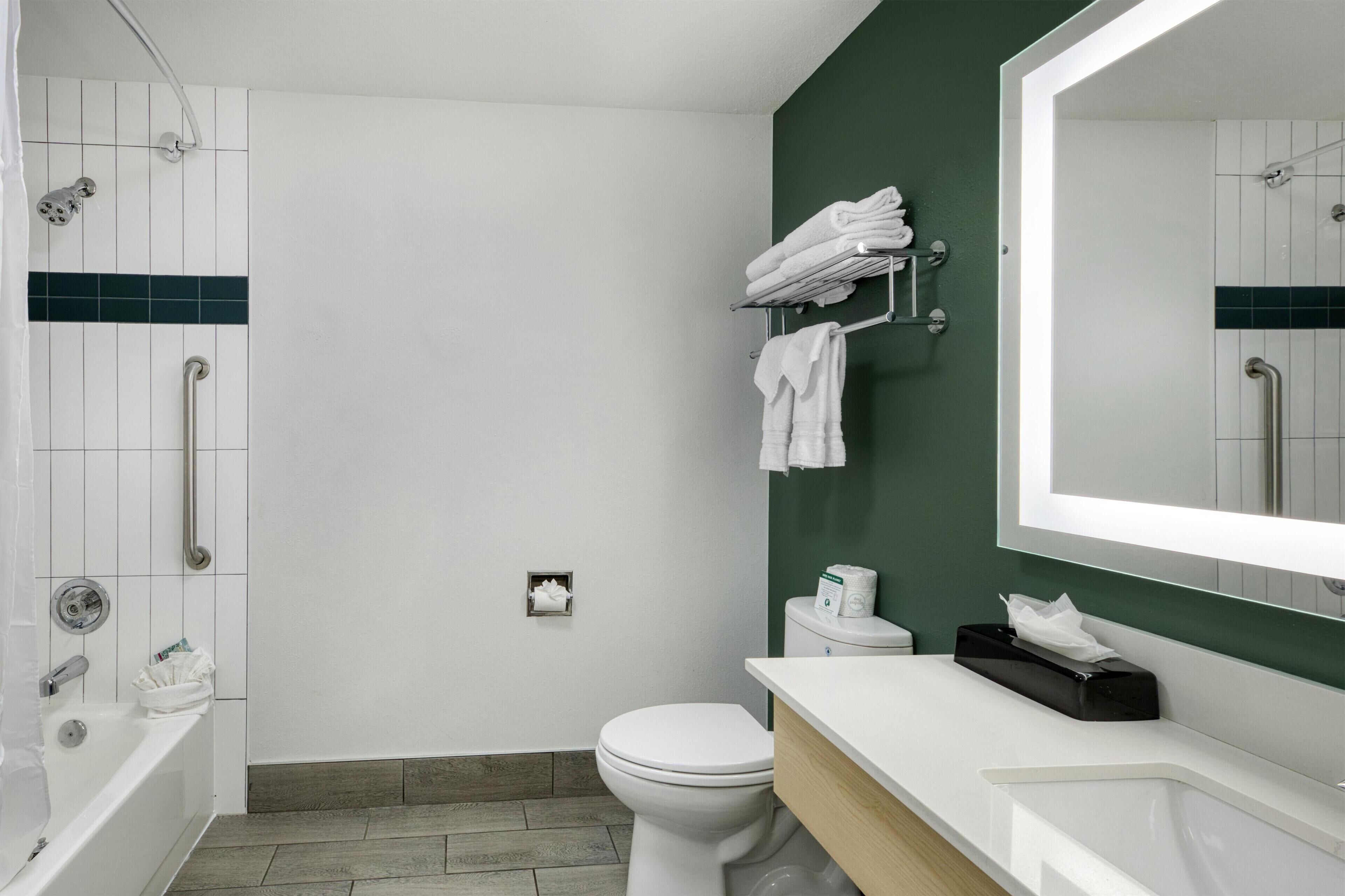 A modern bathroom with a white bathtub, a toilet, and a sink. There is a green accent wall and a large mirror above the sink.