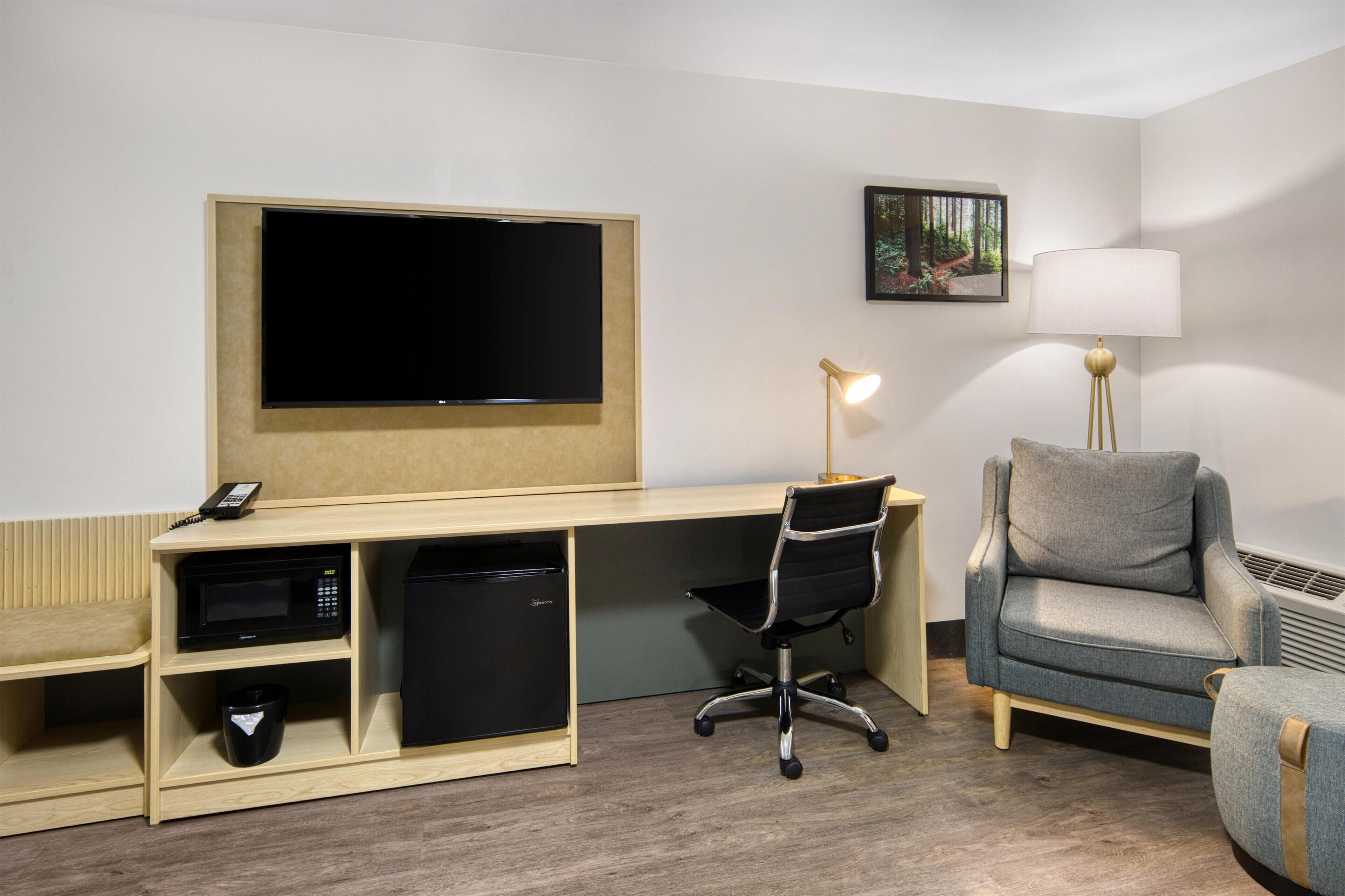 Een hotelkamer met een houten bureau, een zwarte bureaustoel, een grijze bank, een flatscreen televisie, een lamp en een kleine koelkast.
