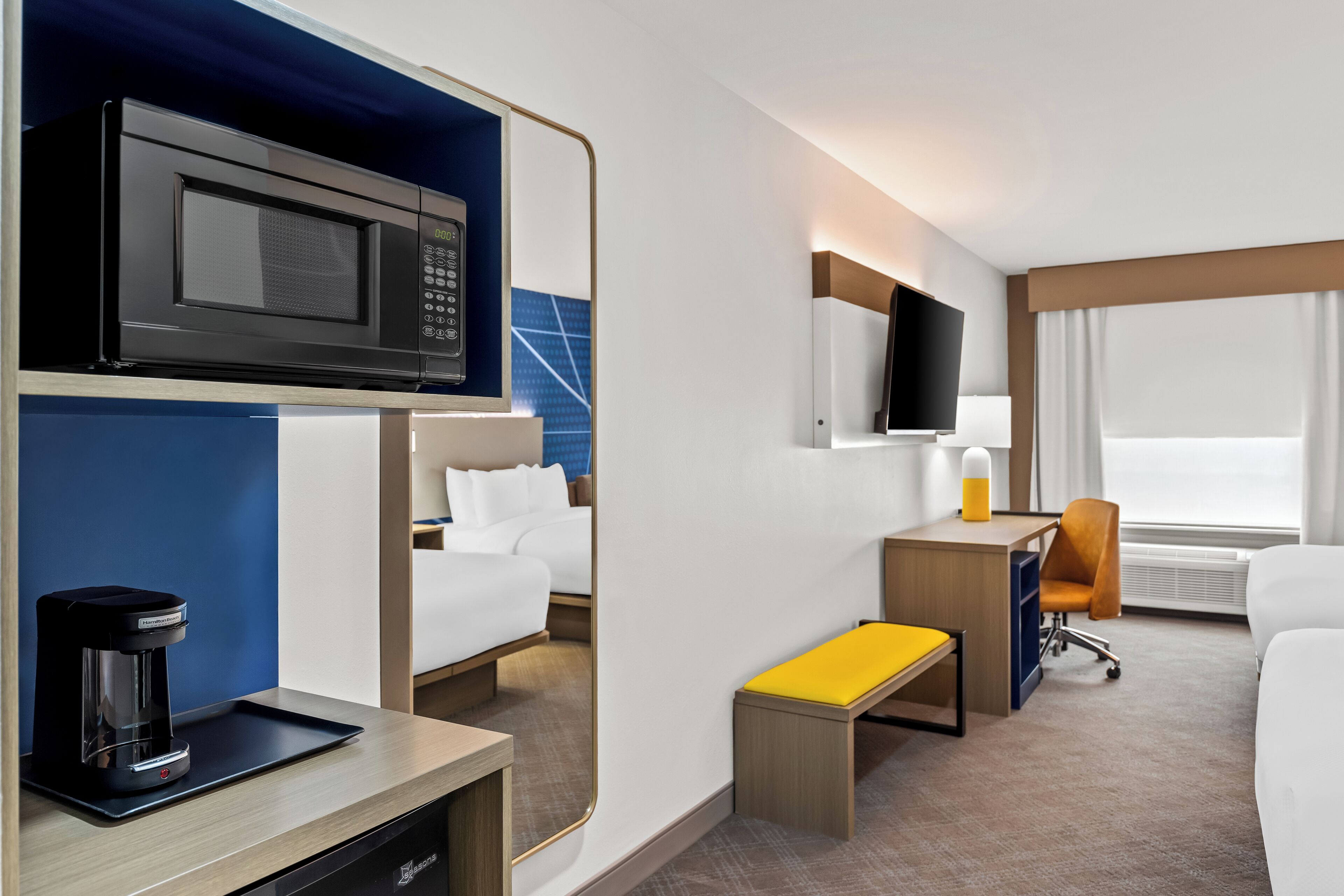 A hotel room with a bed, a desk with a chair, a coffee maker, a television, and a window with curtains.