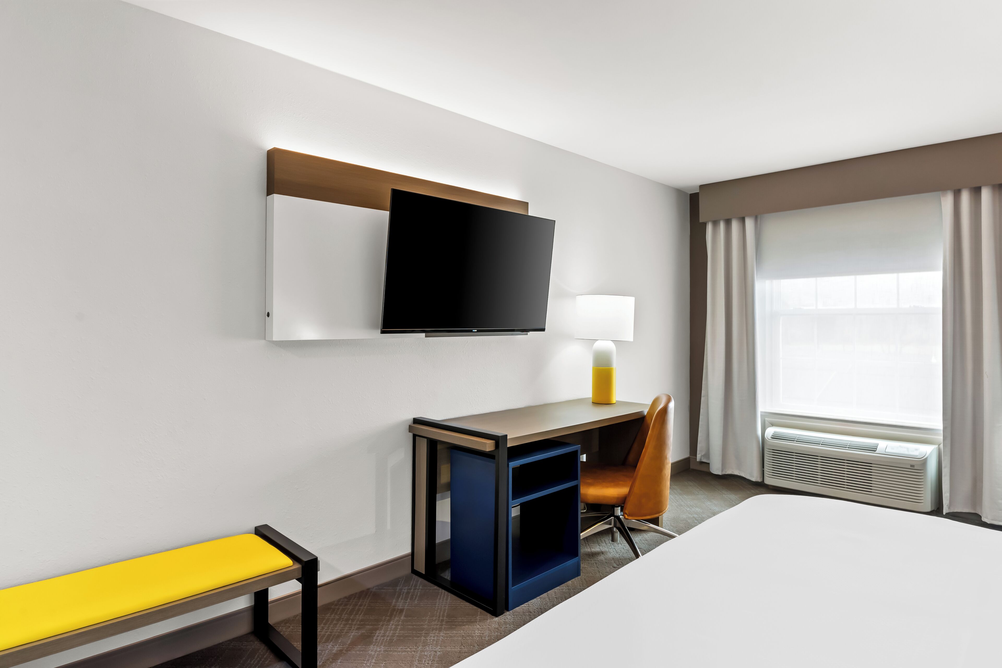 A hotel room with a bed, a desk with a TV, a chair, a bench, and a window with curtains.