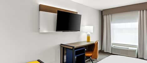 A hotel room with a bed, a desk with a TV, a chair, a bench, and a window with curtains.
