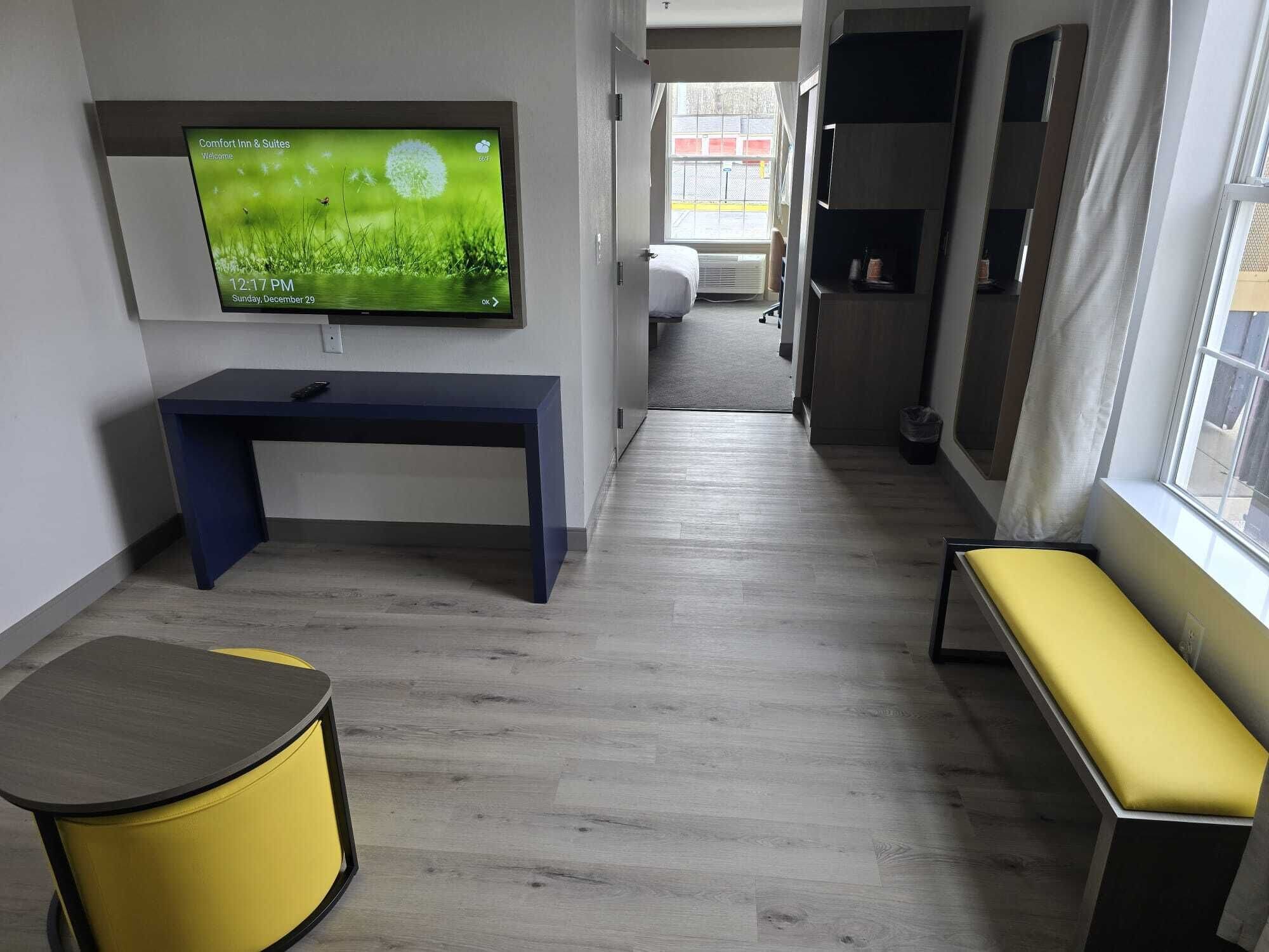 A modern hotel room with a flat-screen TV on the wall, a desk, a bench, and a small round table.