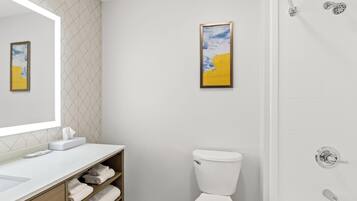 Un baño moderno con un lavamanos blanco, inodoro y ducha. Hay una foto en un marco en la pared y un perchero con toallas.
